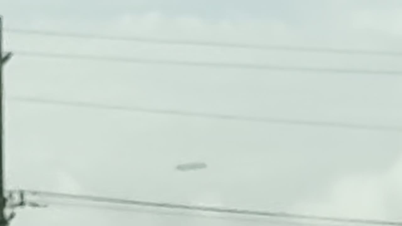 Enlarged frame: a dark elongated object above power lines and a rooftop.